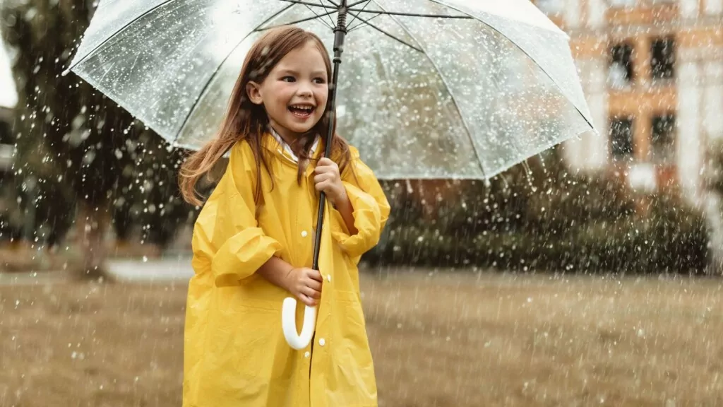 a little girl in a yellow raincoat holding up an umbrella on a rainy day
