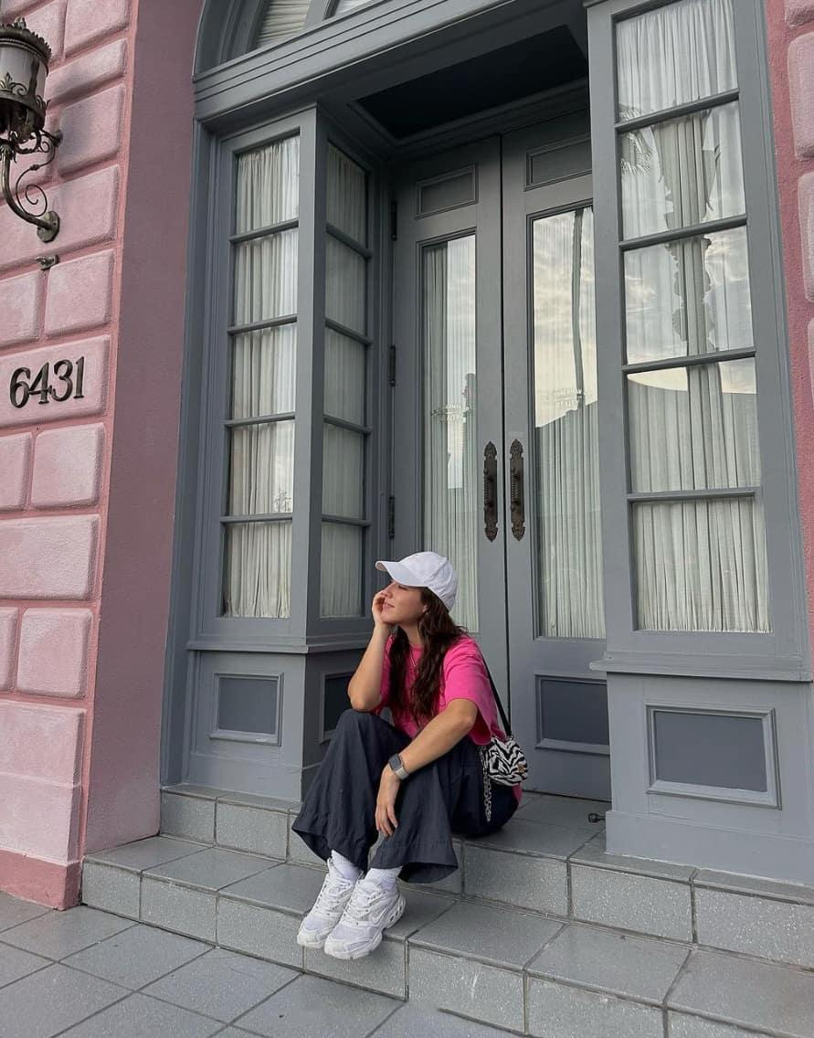 A woman at Universal Studios wearing relaxed black pants, a bright pink tee, chunky white sneakers, and a white baseball cap