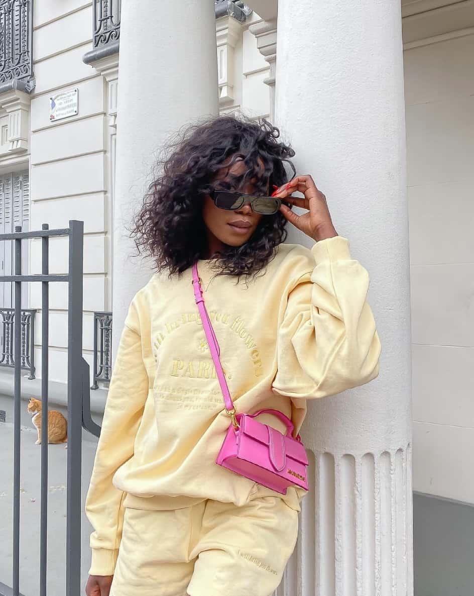 A woman wearing a matching pastel yellow sweatsuit set with black sunglasses and a bright pink handbag