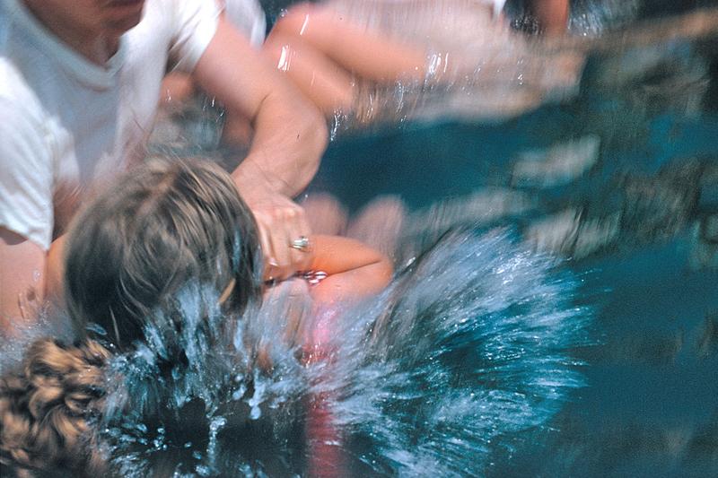 An action shot of a baptism, the person being baptized is blurry, and water splashes around them as they are immersed in the pool by the officiant.