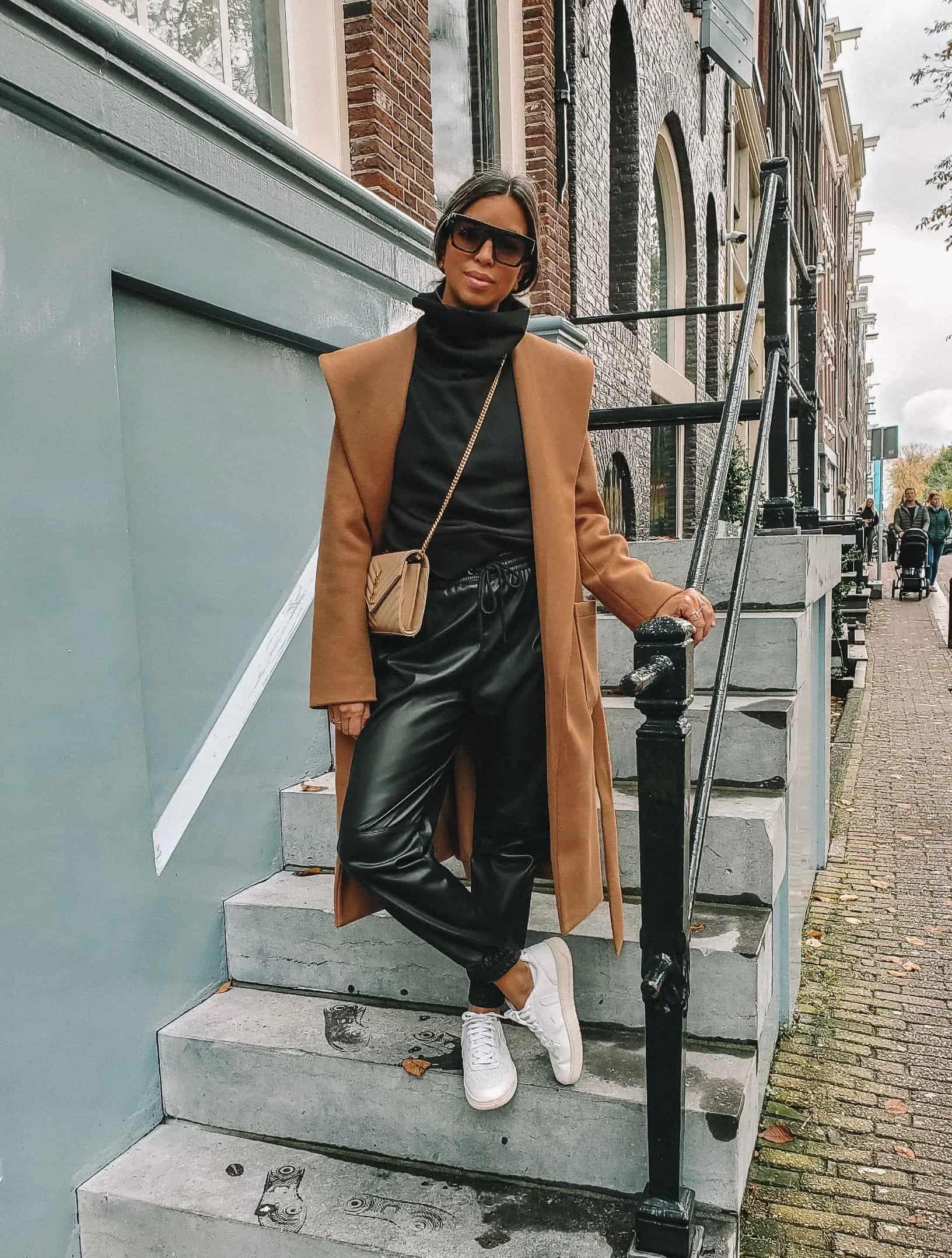 Black woman wearing a long camel coat over a black turtleneck and black leather joggers with white sneakers