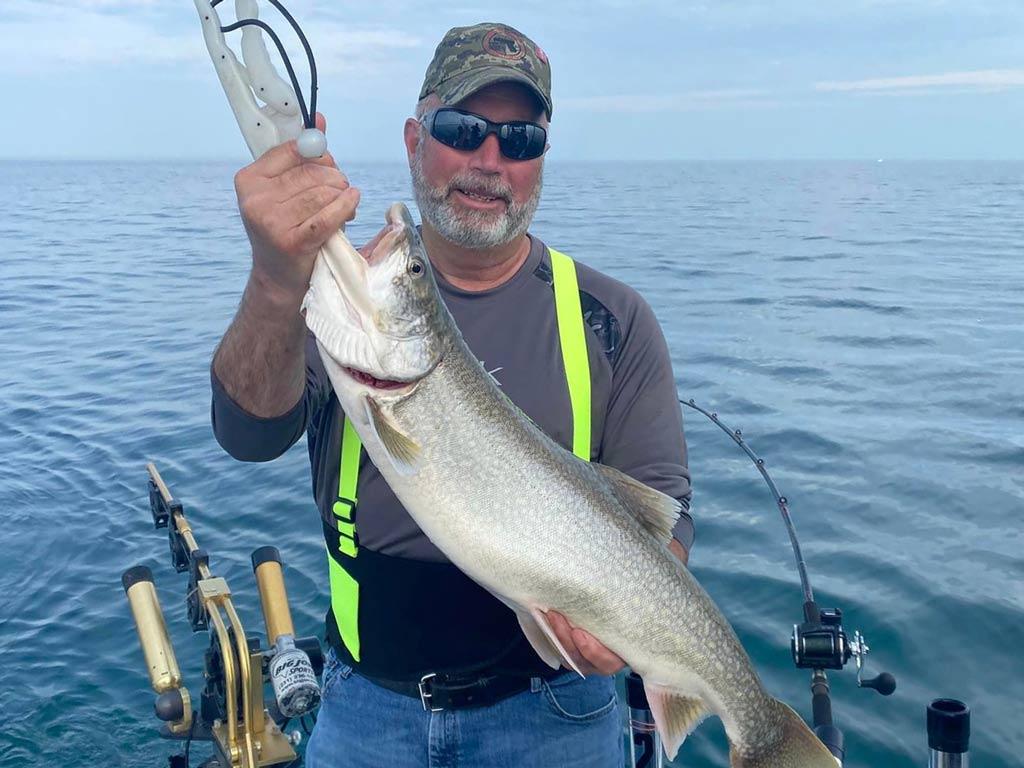 A man on a boat, holding a Lake Trout with fishing gear and Lake Erie