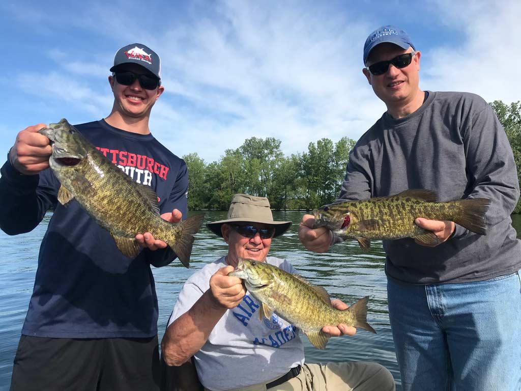 Three anglers on a boat, each holding a big Smallmouth Bass.