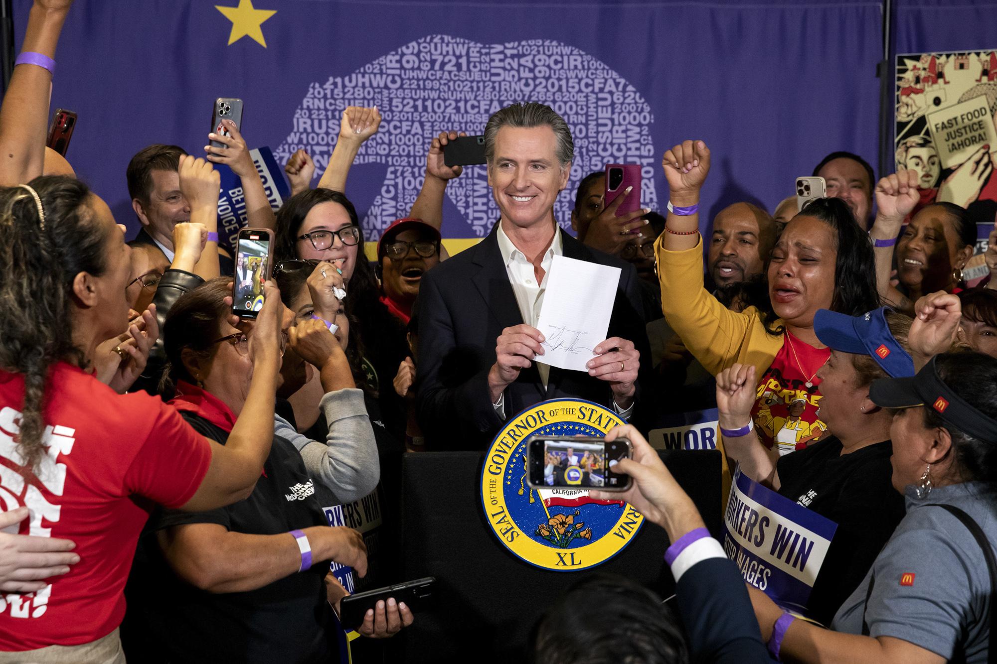 Governor Gavin Newsom poses with cheering workers after he signed legislation supporting fast food workers and boosting wages to $20 an hour, at a press conference at SEIU Local 721 in Los Angeles on Sept. 28, 2023. Anneisha Williams, a fast food worker cheers beside him to the right. Photo by Alisha Jucevic for CalMatters