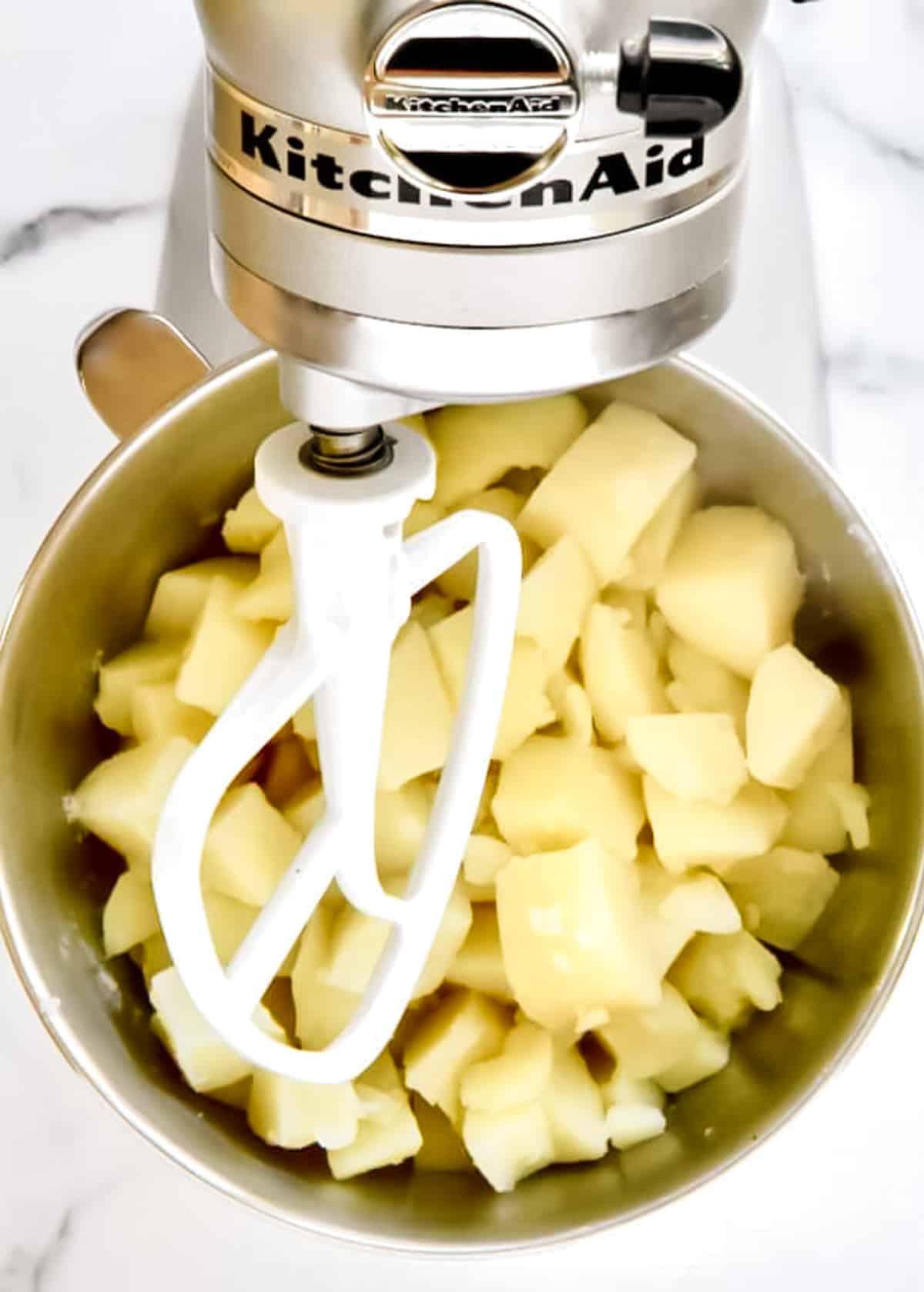 The bowl of a kitchenaid mixer fitted with a paddle attachment. The bowl contains cooked potatoes, not yet mashed.