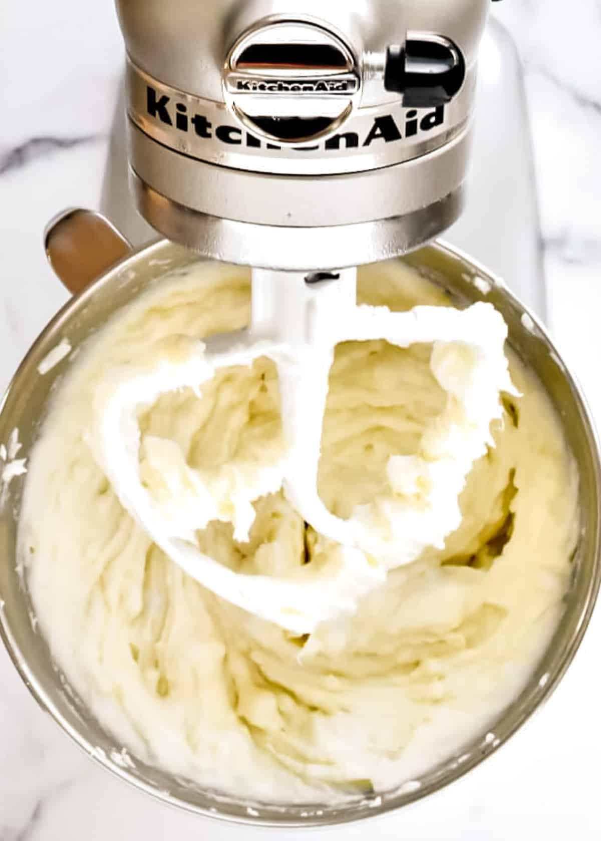 The bowl of a kitchenaid mixer fitted with a paddle attachment. The bowl contains cooked and mashed potatoes.