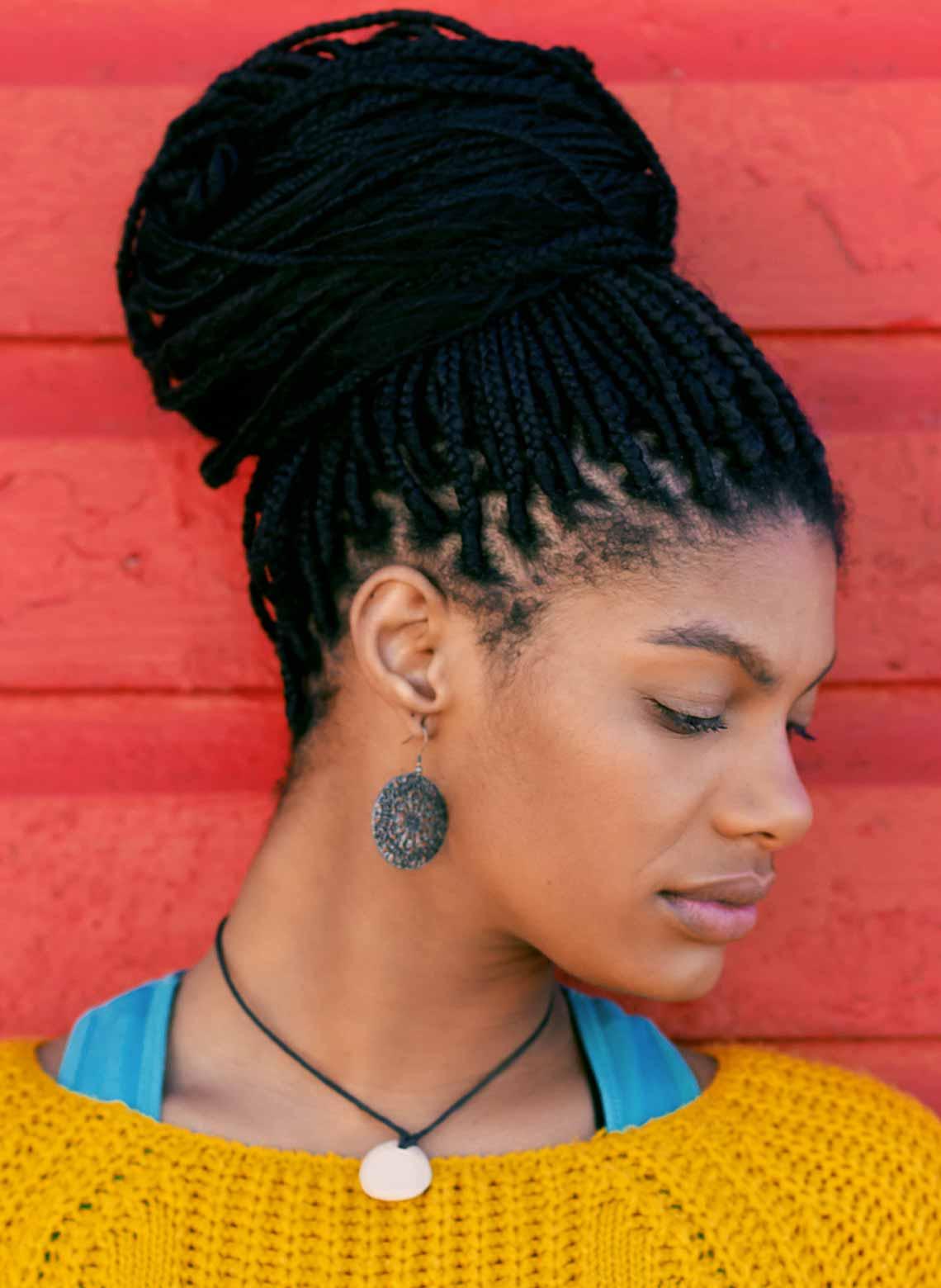 person wearing a yellow knitted top, light blue tank straps showing around the neck, a circular pendant and earrings, and small box braids tied up in a bun