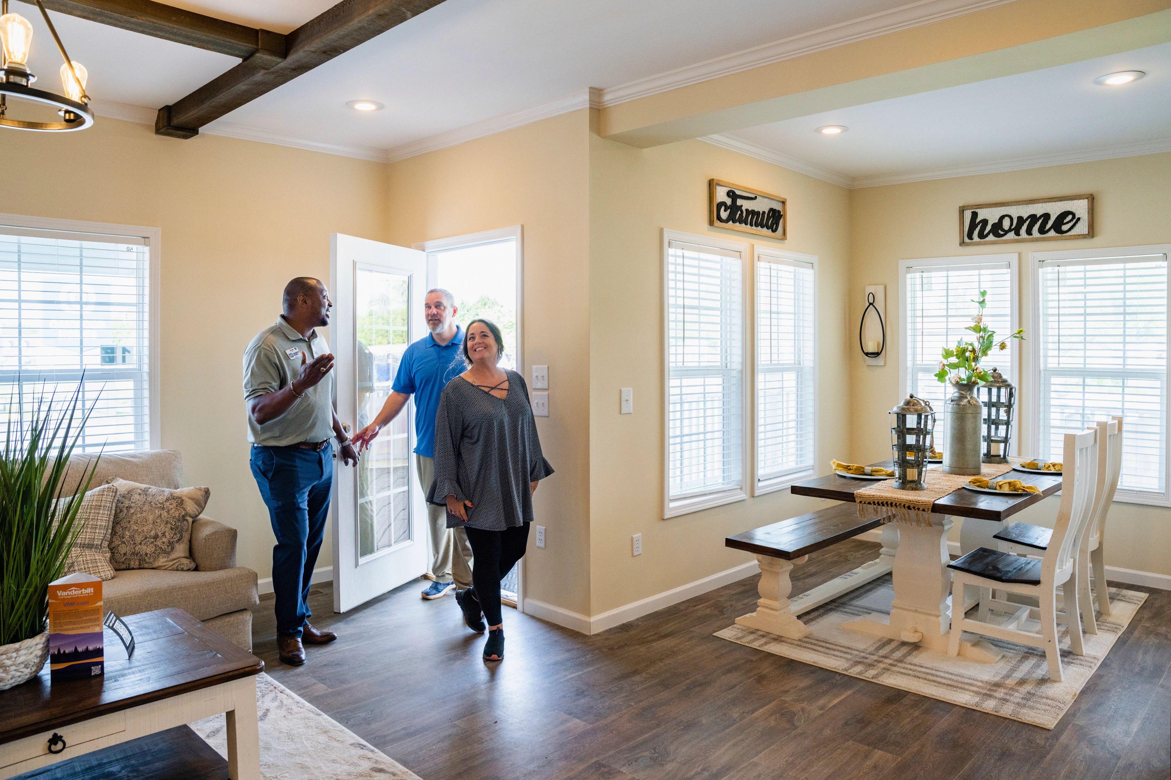A couple walks through an open door of a manufactured home while a home consultant gestures for them to look around.