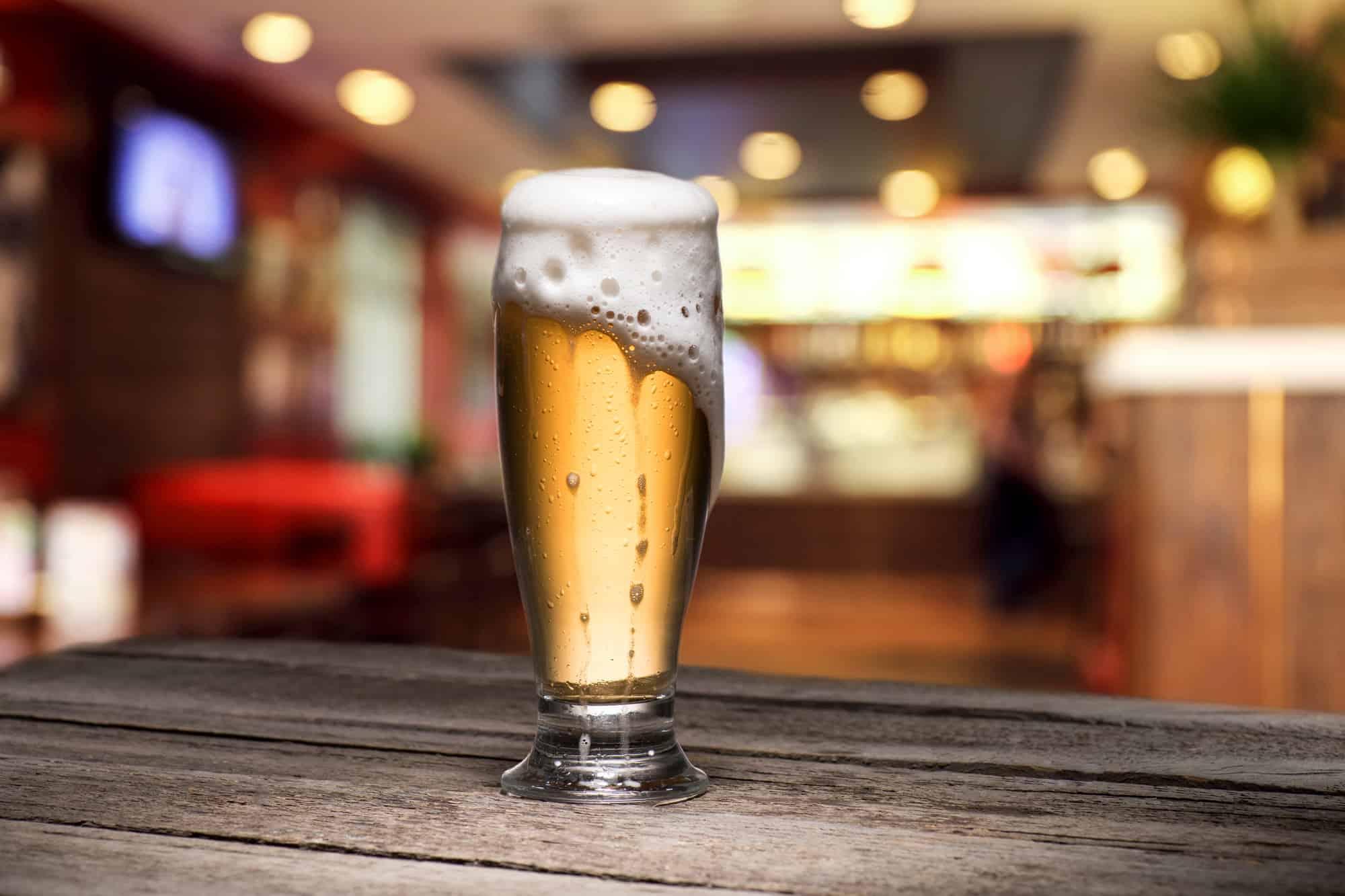 a tall beer glass full of beer with foam overflowing down the side of the glass