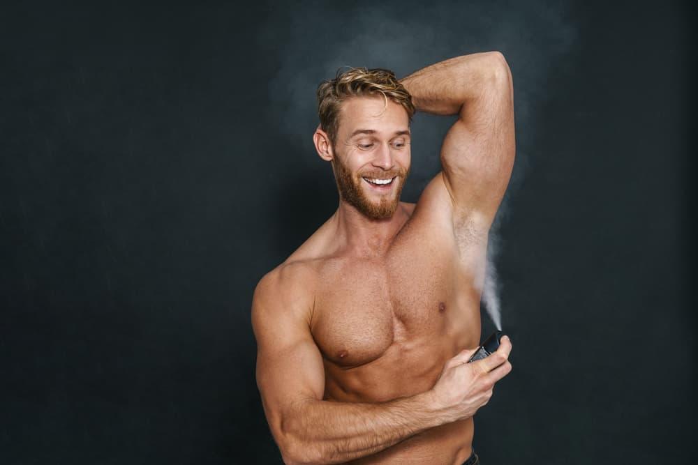Happy caucasian shirtless man smiling while using deodorant isolated over black background
