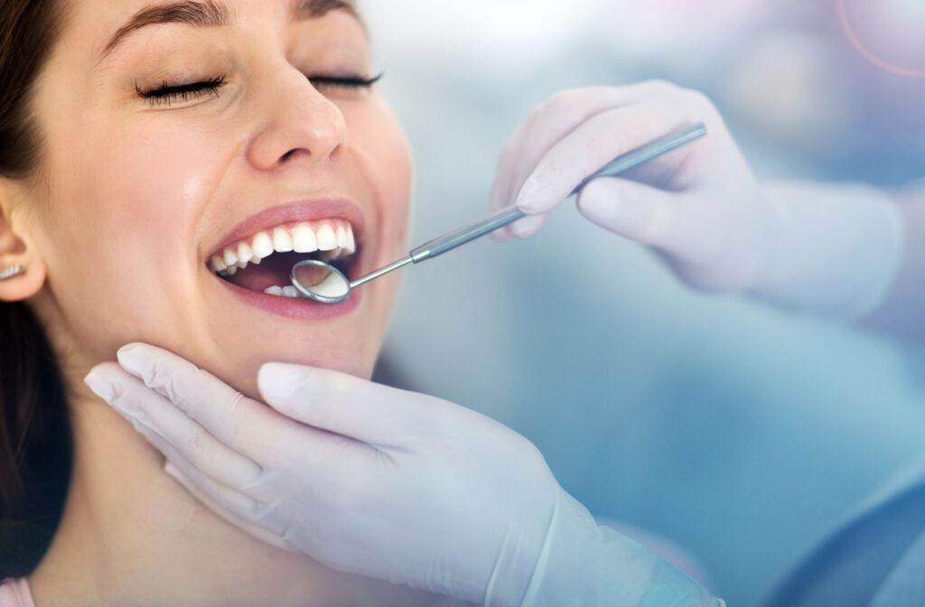 A woman smiles with her eyes closed while getting her teeth examined by a dentist with a small mirror