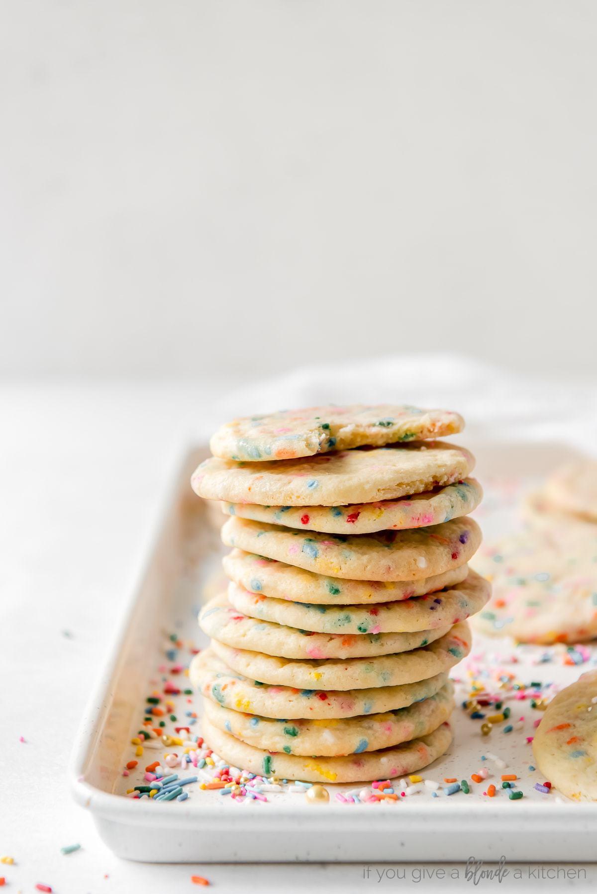 stack of a dozen sprinkle sugar cookies on a baking sheet covered in rainbow sprinkles