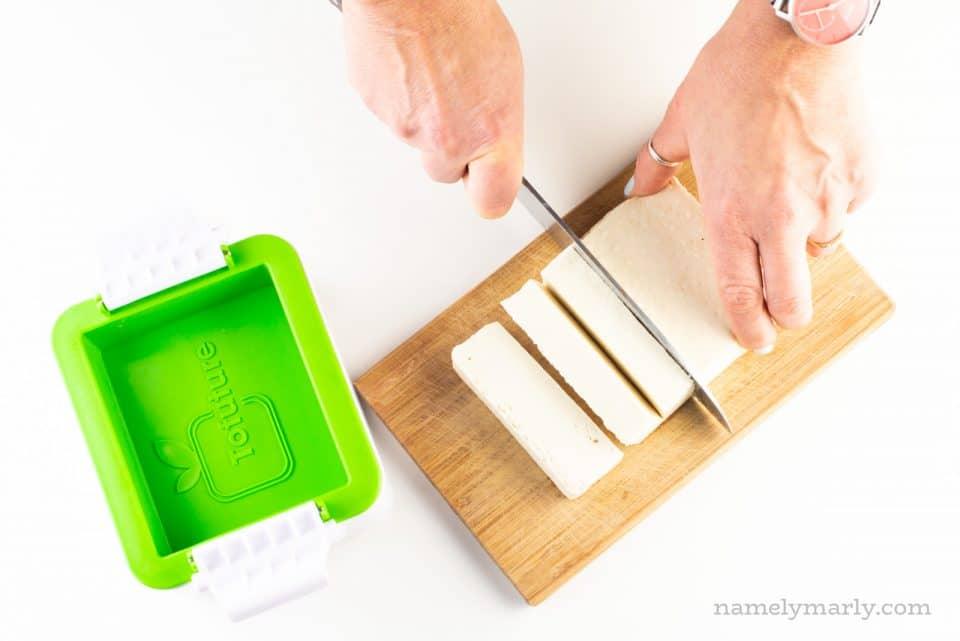 A hand holds a knife and is cutting tofu. It