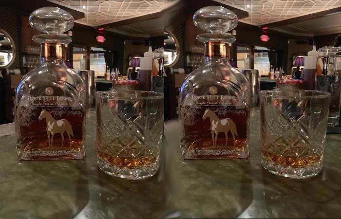 A bottle of Rock Hills Farms Bourbon with a glass of bourbon sits on a green bar top