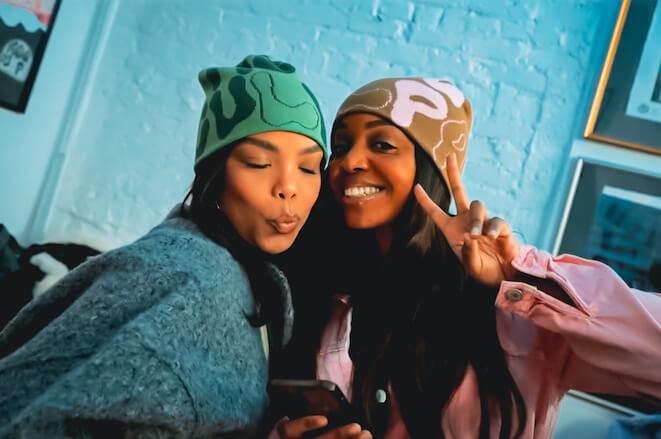Two women wearing Mea Culpa beanies post for the camera at the pop-up shop celebration. One has eyes closed and is making kissy lips. The other, smiling, flashes a peace sign.
