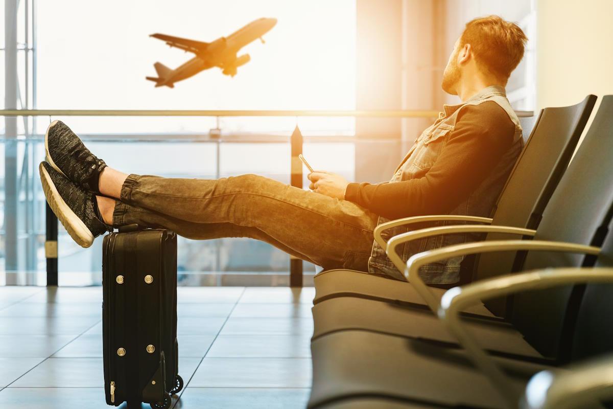 Man sat in an airport waiting area with his legs up on a suitcase, looking out at a plane taking off outside of the window